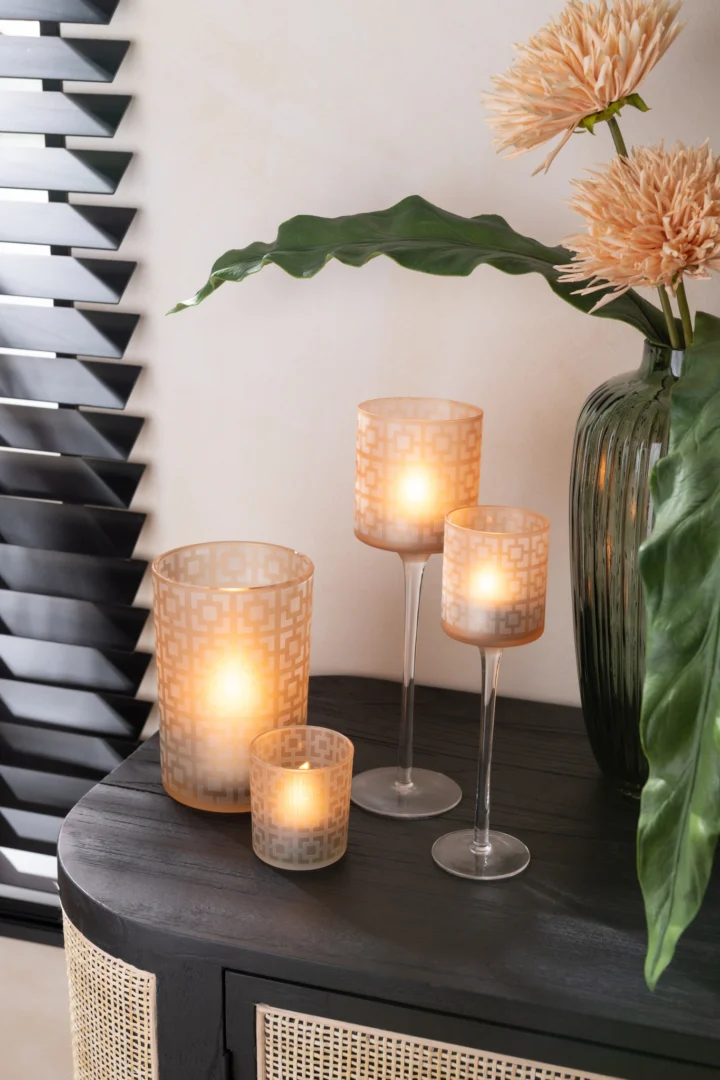 Een zwarte tafel met drie brandende Theelichthouder Vierkant Glas Beige Klein kandelaars van verschillende hoogtes naast een groene glazen vaas gevuld met roze bloemen en grote groene bladeren. Zwarte jaloezieën bevinden zich op de achtergrond.