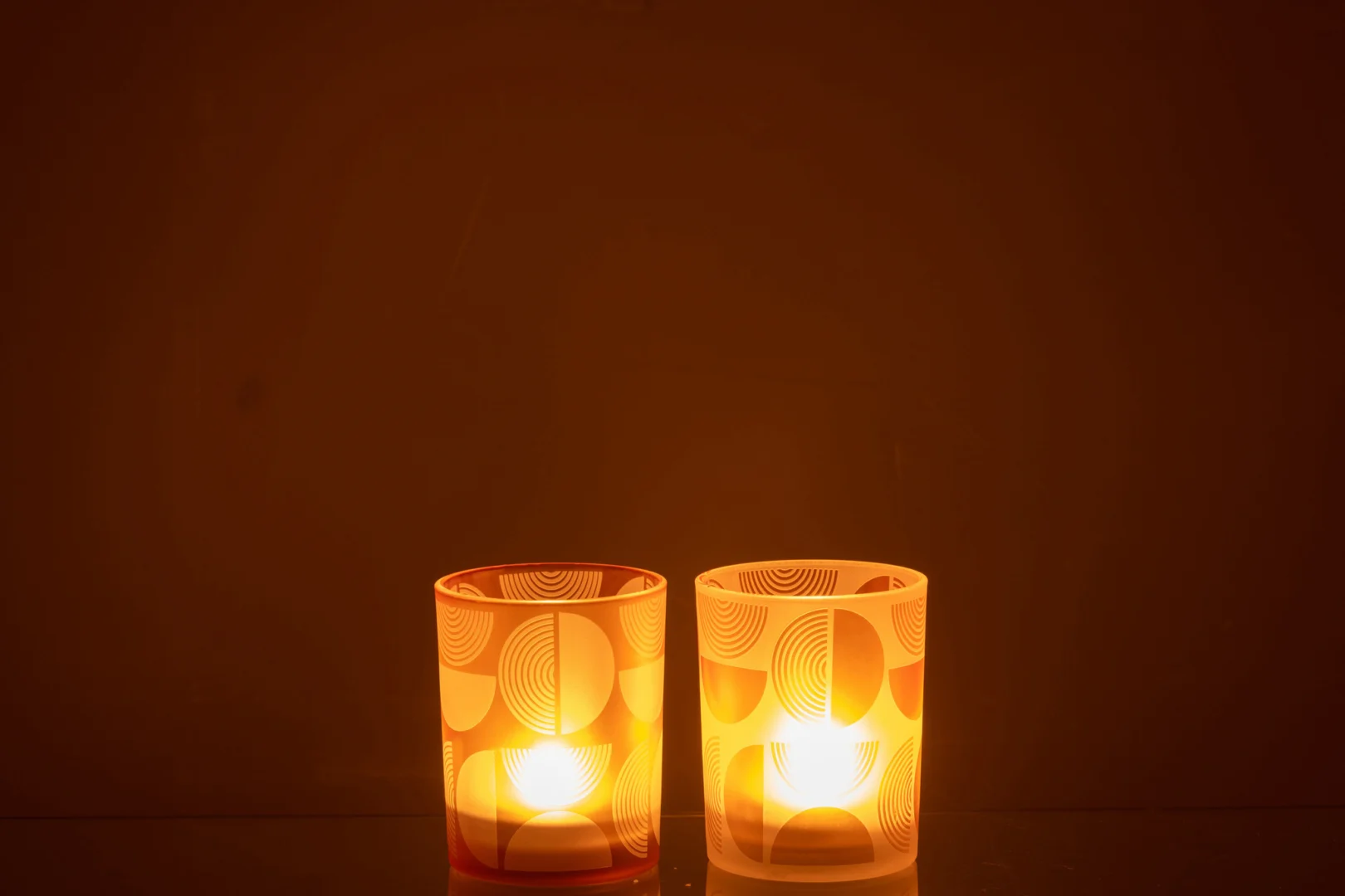 Twee Orkaan Halve Cirkels Glas Oranje Medium kaarsen, in houders met geometrische motieven, zitten naast elkaar tegen een donkerbruine achtergrond en werpen een warme en gezellige gloed. Dit assortiment van 2 kaarsen creëert een vredige en uitnodigende sfeer.