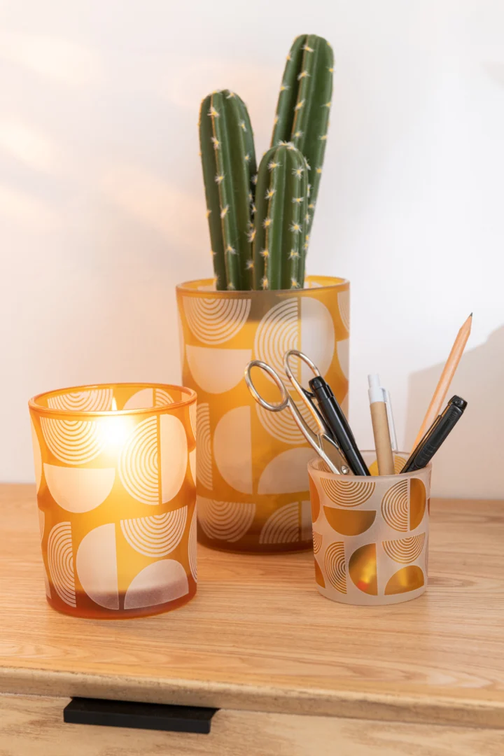 Twee Orkaan Halve Cirkels Glas Oranje Medium containers staan uitgestald op een houten oppervlak; in de ene zit een cactus, in de andere pennen en een schaar.