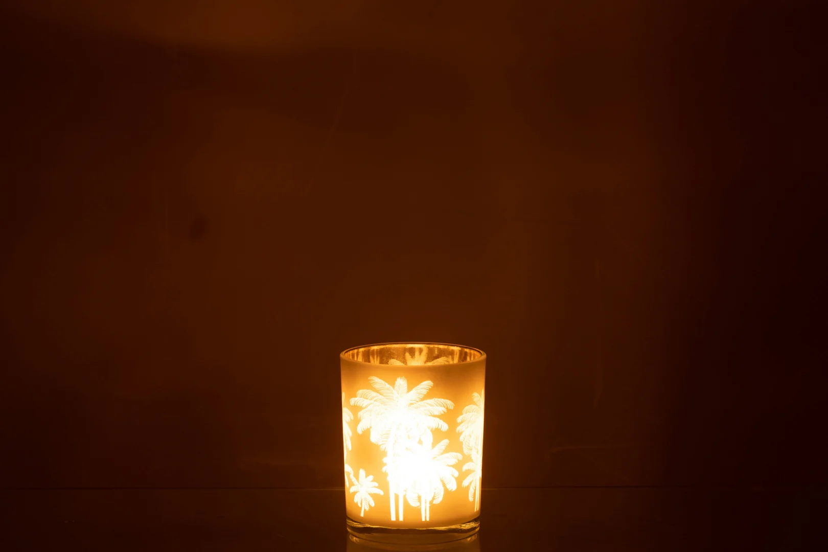 De Kaarsenhouder Orkaan Palm Bomen Glas Blauw Goud Medium werpt een warme gloed, met palmboomsilhouetten die door de glazen houder tegen een donkere achtergrond schijnen.