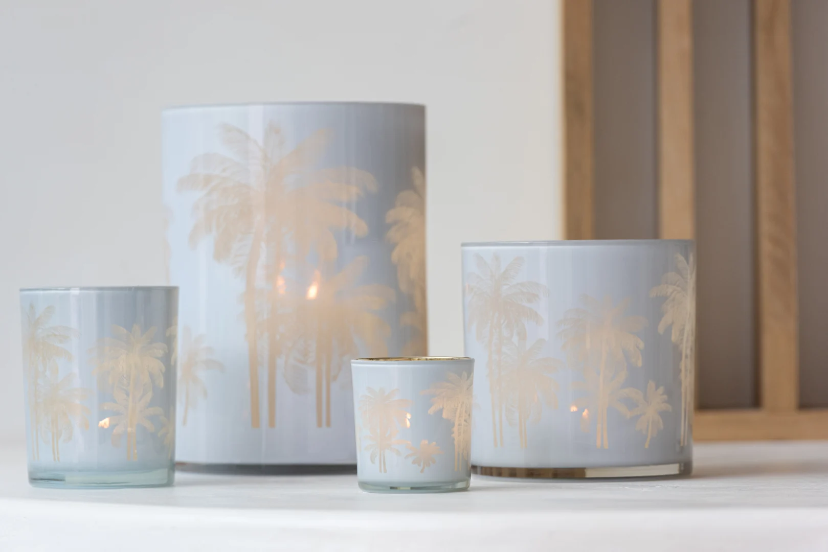 Vier Kaarsenhouder Orkaan Palm Bomen Glas Blauw Goud Medium kandelaars, met geëtste palmboommotieven, zijn gerangschikt op een wit oppervlak. Brandende kaarsen binnenin werpen een warme gloed door het semi-transparante blauwe en gouden glas.