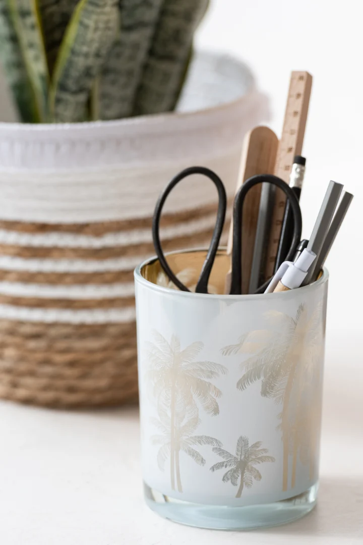 De Kaarsenhouder Orkaan Palm Bomen Glas Blauw Goud Medium bevat een schaar, pennen, een potlood en een liniaal. Op de achtergrond staat een groene potplant in een gevlochten mand.