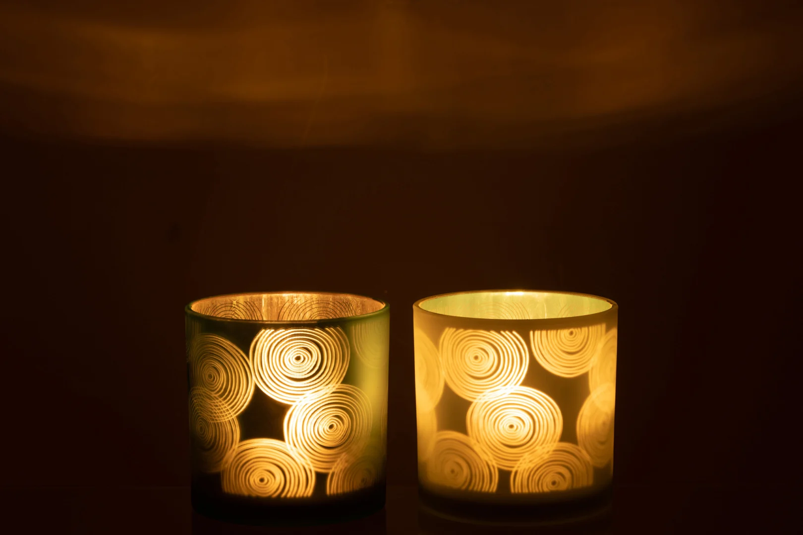 De Orkaan Spiralen Glas Mix Groot Assortiment Van 2 heeft twee glazen kaarsen met spiraalpatronen, die een warme gele gloed uitstralen en zacht licht en wervelende patronen werpen tegen een donkere achtergrond.