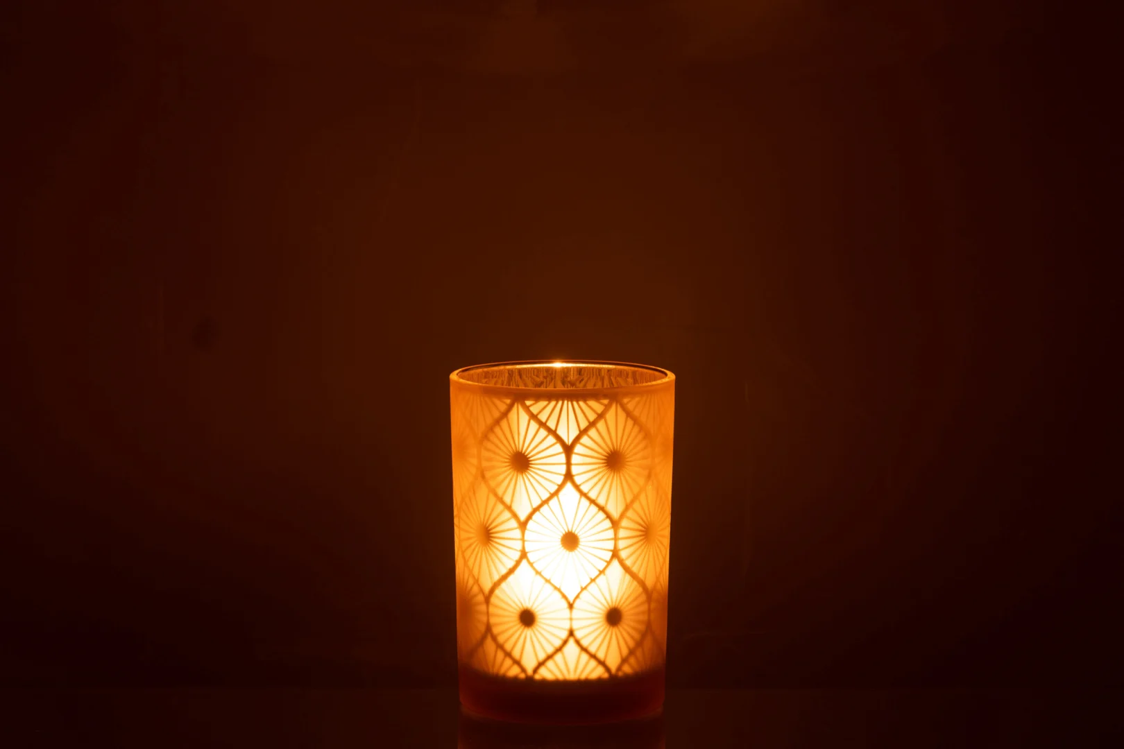 De Kaarsenhouder Orkaan Rhomb Bloemen Glas Geel Groot werpt een warme, zachte gloed en ingewikkelde schaduwen vanuit de glazen houder met patroon en creëert zo een gezellige sfeer tegen een donkere achtergrond.