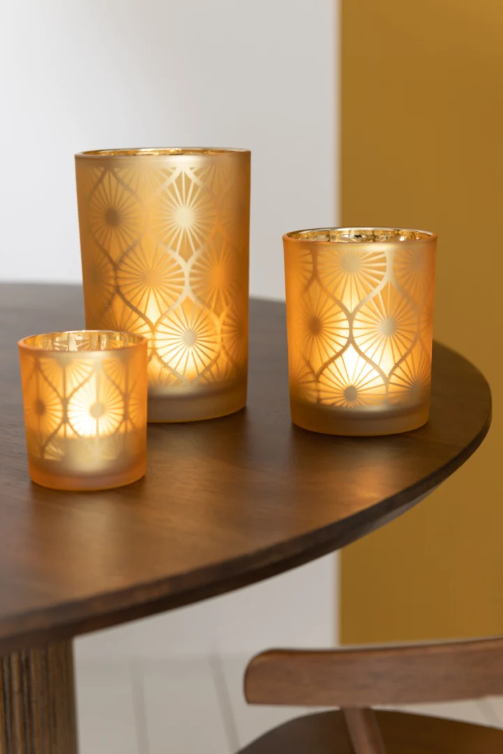 De Kaarsenhouder Orkaan Rhomb Bloemen Glas Geel Groot, met een brandende kaars erin, staat op een ronde houten tafel en werpt een warme gloed. Het tafereel speelt zich af tegen witte en mosterdgele muren.