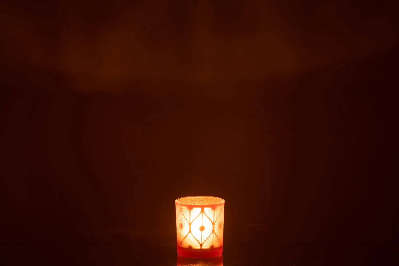 De Theelichthouder Ruit Bloemen Glas Oranje Klein, met één brandende kaars erin, gloeit zacht in het midden van de donkere kamer en werpt warm licht en zachte schaduwen rondom.