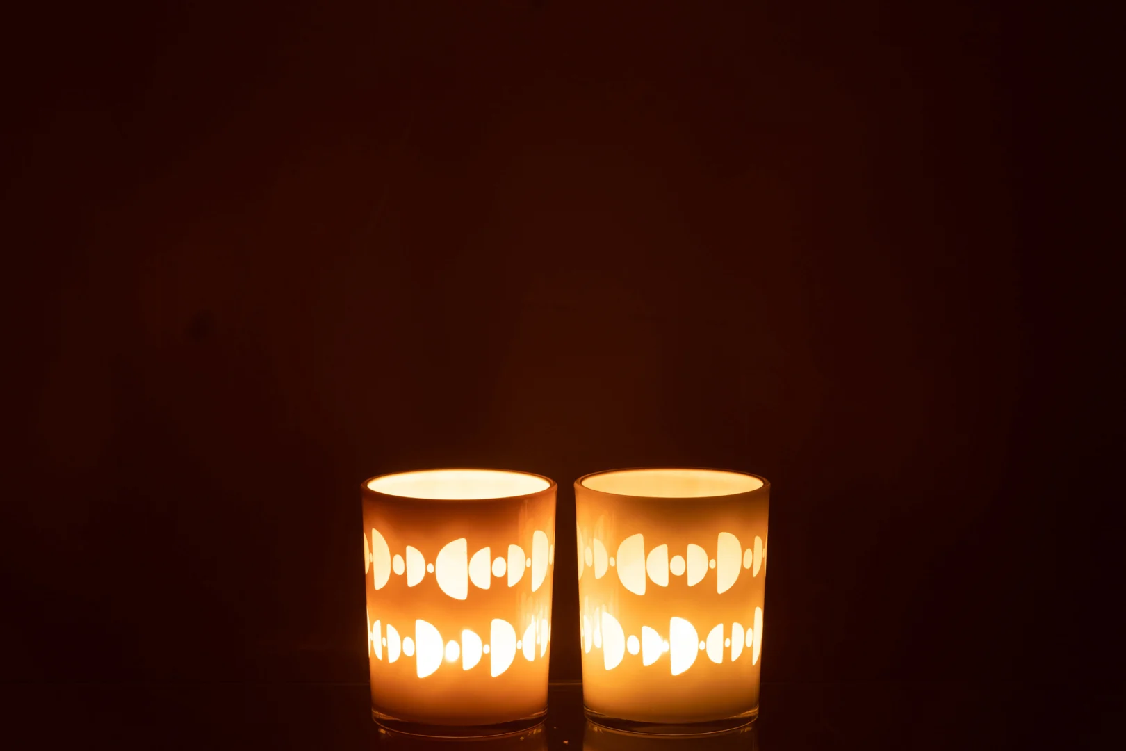 De Orkaan Half Moon Glass Mix Medium Assortiment Van 2 heeft twee brandende kaarsen in glazen houders met halvemaanvormige en ovale uitsparingen, die een warm, gloeiend licht werpen tegen een donkere achtergrond.