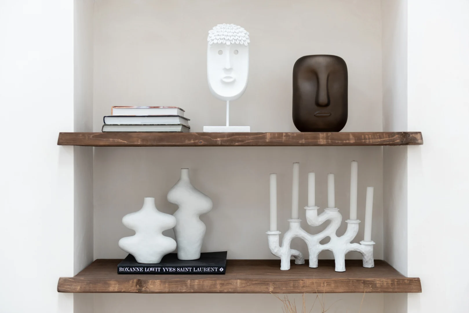 Twee houten planken tonen abstracte gezichtssculpturen, gestapelde boeken, twee witte vazen en de Kaarsenhouder Hars Wit 6 Armig kandelaar. Een witte muurachtergrond versterkt de moderne minimalistische stijl.