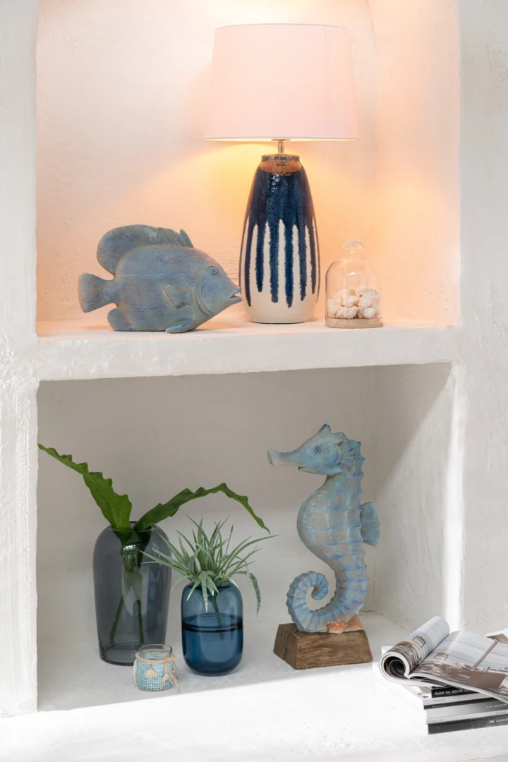 Een witte alkoofplank toont een blauwe lamp met een beeld van een vis, een pot met schelpen, een zeepaardje, twee glazen vazen met groene planten, Theelichthouder Zeedecoratie Glas Blauw/Wit en een opgerold tijdschrift.