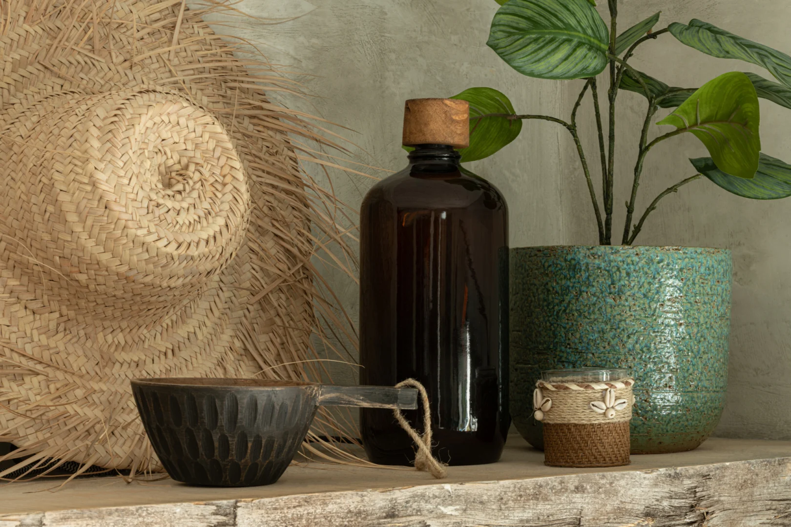 De Theelichthouder Schelpen Glas Bruin staat op een rustieke houten plank naast een strooien hoed, zwarte keramische beker, decoratieve container en groene potplant, allemaal tegen een grijze muur met structuur.