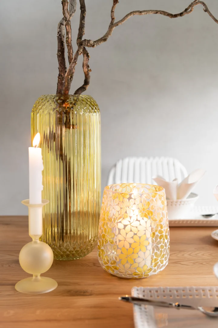 Een houten gedekte tafel met de Kaarsenhouder Orb Glas Lichtgeel Klein Set 2, met gloeiende kaarsen, naast een brandende witte kaars in een matte houder en wit servies, met stoelen op de achtergrond.