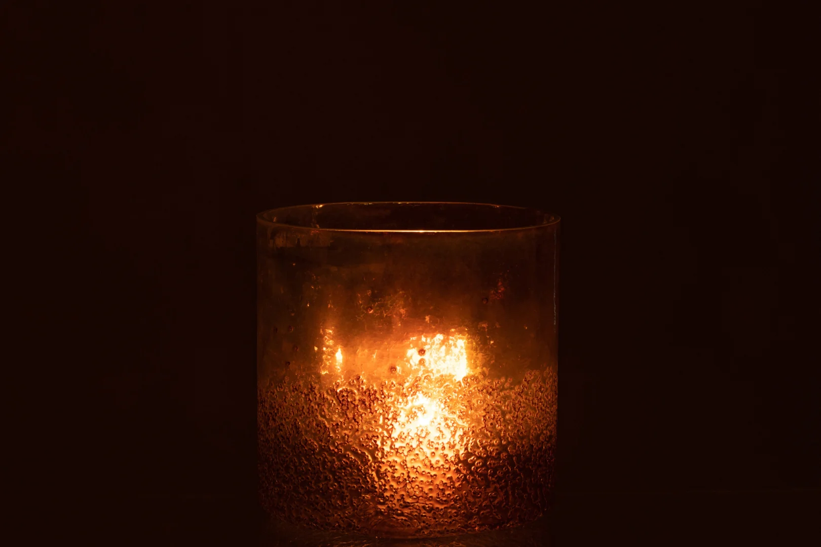 De Hurricane Glas Bruin Groot gloeit warm met een brandende kaars erin, de glazen houder met textuur werpt een zacht amberkleurig licht tegen een donkere achtergrond.