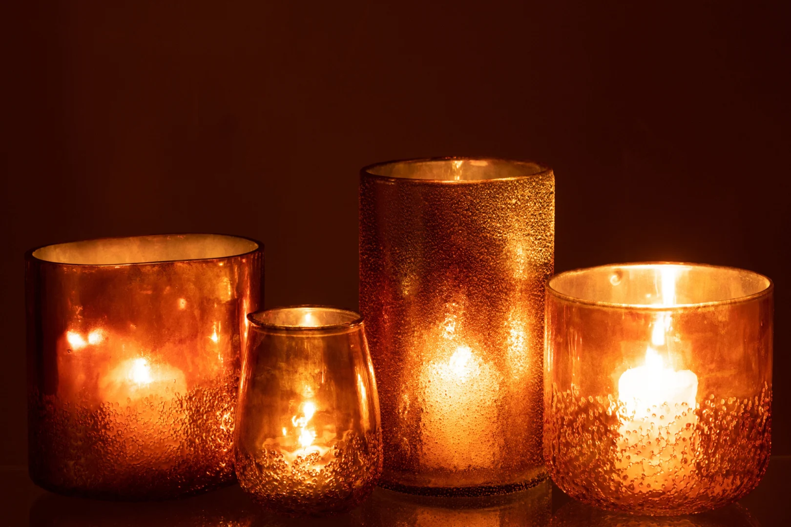 Vier Spiegel Goud Glas Extra Groot Formaat kaarsen in gestructureerde amberkleurige glazen houders van verschillende hoogtes stralen een warme, zachte gloed uit tegen een donkere achtergrond.