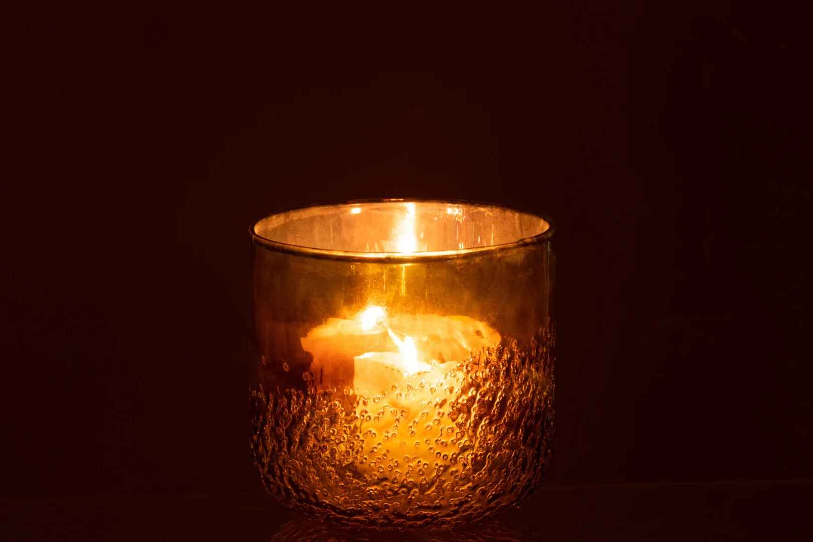 De Spiegel Groen Glas Medium Formaat kandelaar gloeit warm, het groen getextureerde glas werpt zacht oranje en geel licht in de duisternis.