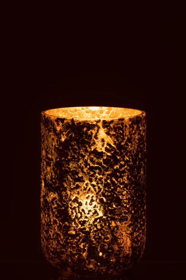 De Kaarsenhouder Orkaan Koper Geoxideerd Groot gloeit warm met een brandende kaars erin, de metalen houder met textuur werpt opvallende patronen van licht en schaduw in een donkere omgeving.