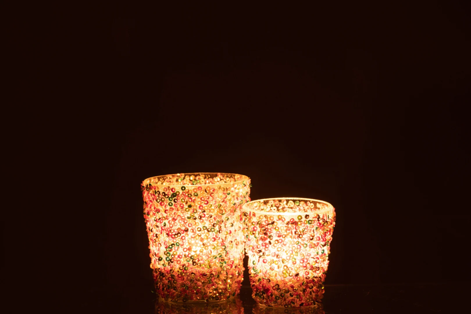 De Kaarsenhouder Orkaan Sequin Glas Pastel Groot heeft kleurrijke kralen die warm oplichten in het donker en een zacht, gezellig licht werpen tegen een effen achtergrond.