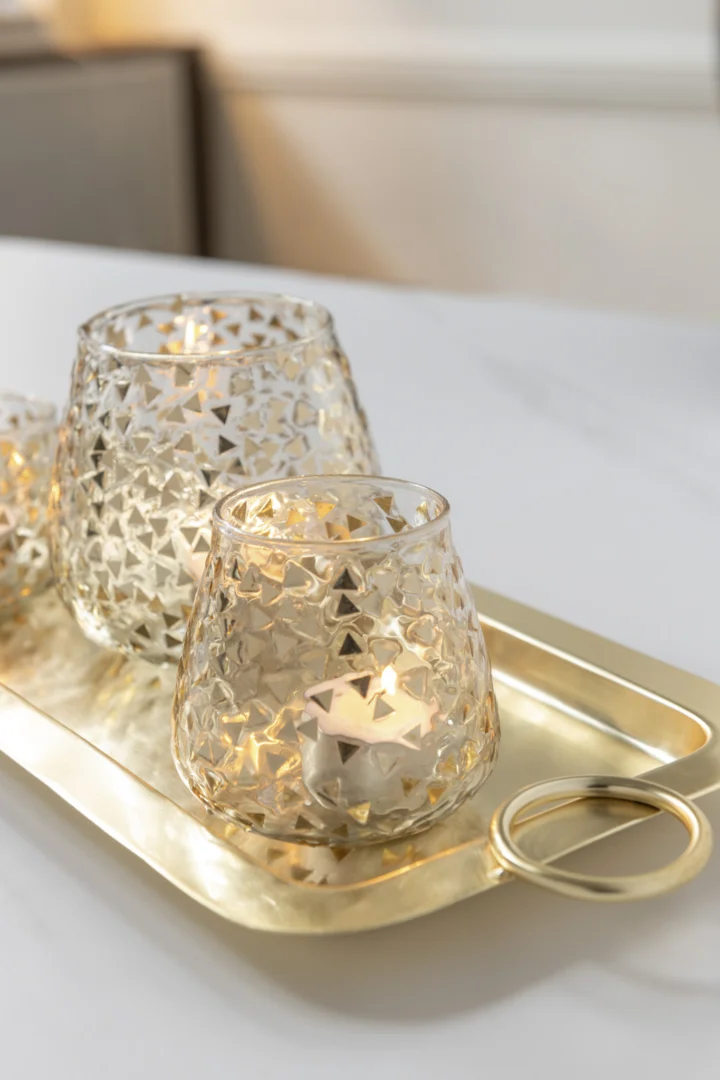 De Kaarsenhouder Orkaan Driehoek Glas Goud Groot, met kaarsen erin, staat op een glanzend gouden dienblad op een wit oppervlak, wat een warme en elegante sfeer creëert.
