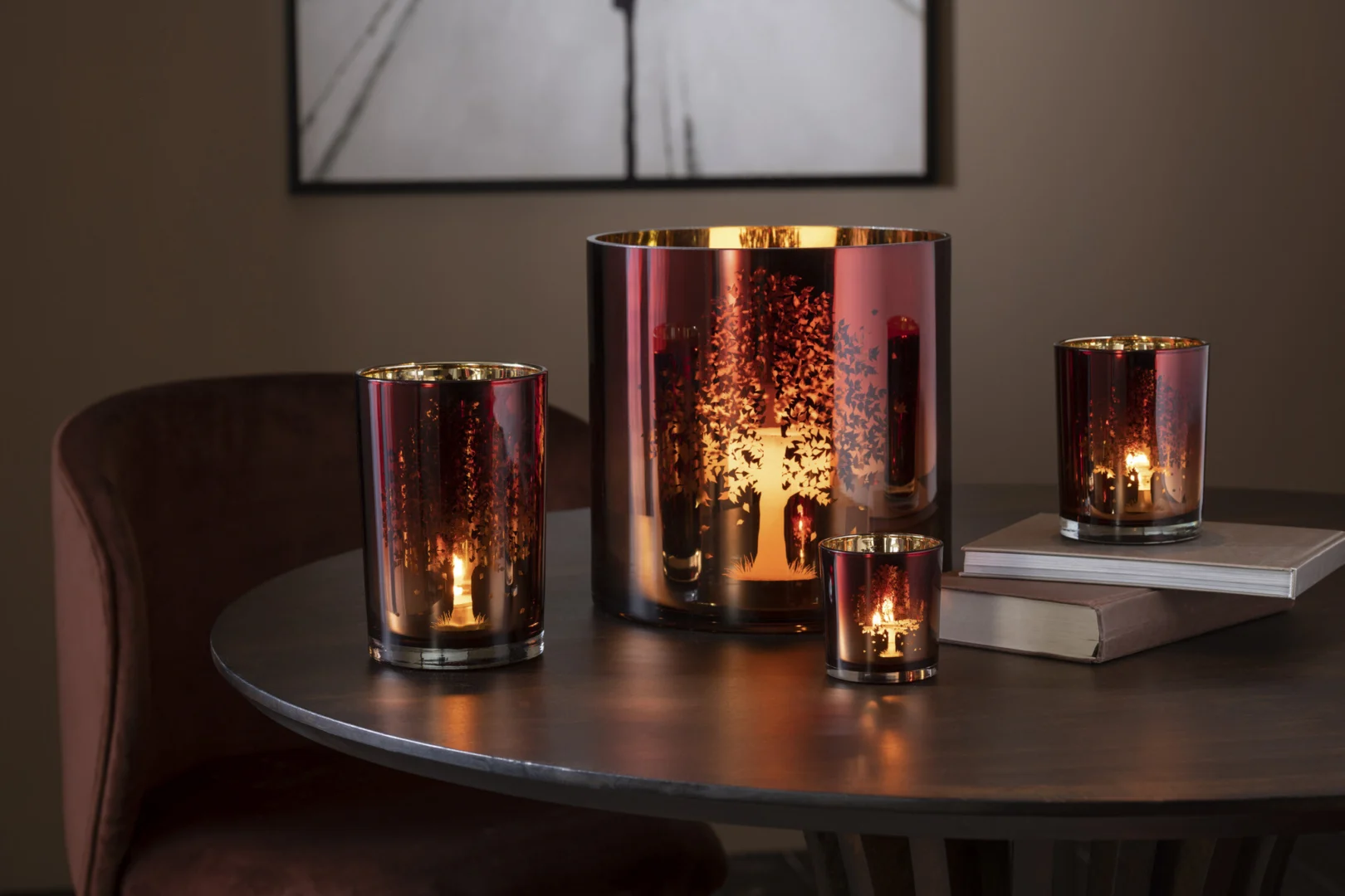 Vier Theelichthouder Bomen Glas Bruin Klein, metalen roodbruine glazen kandelaars van verschillende grootte, met brandende kaarsen erin staan op een ronde donkere tafel naast twee gestapelde boeken en een vage wandklok op de achtergrond.