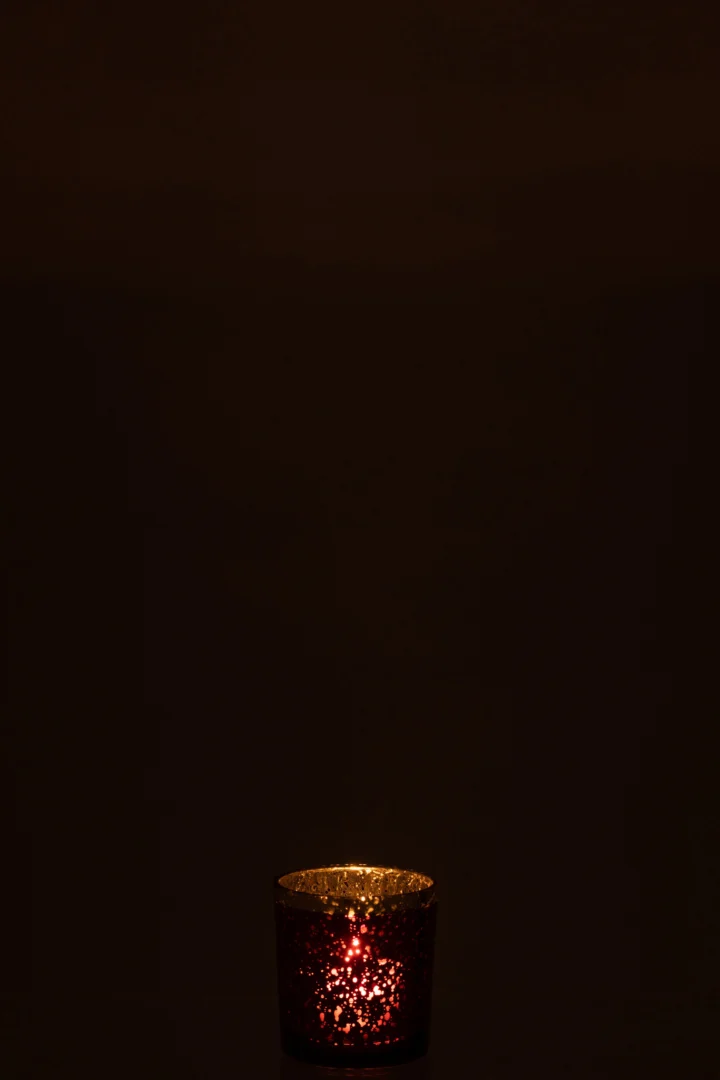 De Theelichthouder Gouden Rand Glas Rood Klein gloeit zacht in het donker en werpt een warm, goudrood licht uit de kleine glazen houder met een gouden rand tegen een zwarte achtergrond.
