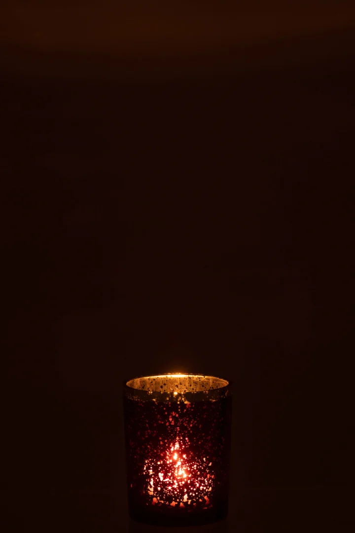 De Kaarsenhouder Orkaan Glas Rood Gouden Rand Medium gloeit zacht en werpt een warm licht uit de rode, gespikkelde glazen houder met een gouden rand tegen een donkere achtergrond.