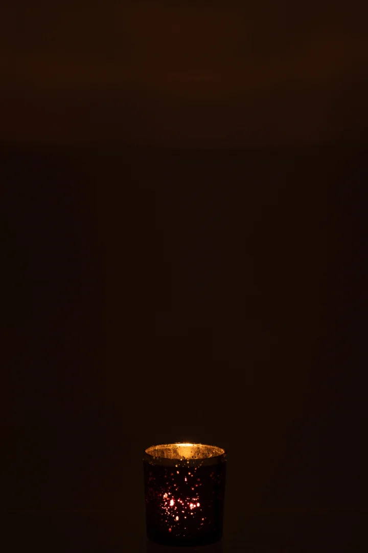 Een enkele brandende kaars gloeit zacht in het donker en werpt een zacht licht door de Theelichthouder met gouden rand glas bordeauxrood klein onderaan de afbeelding.
