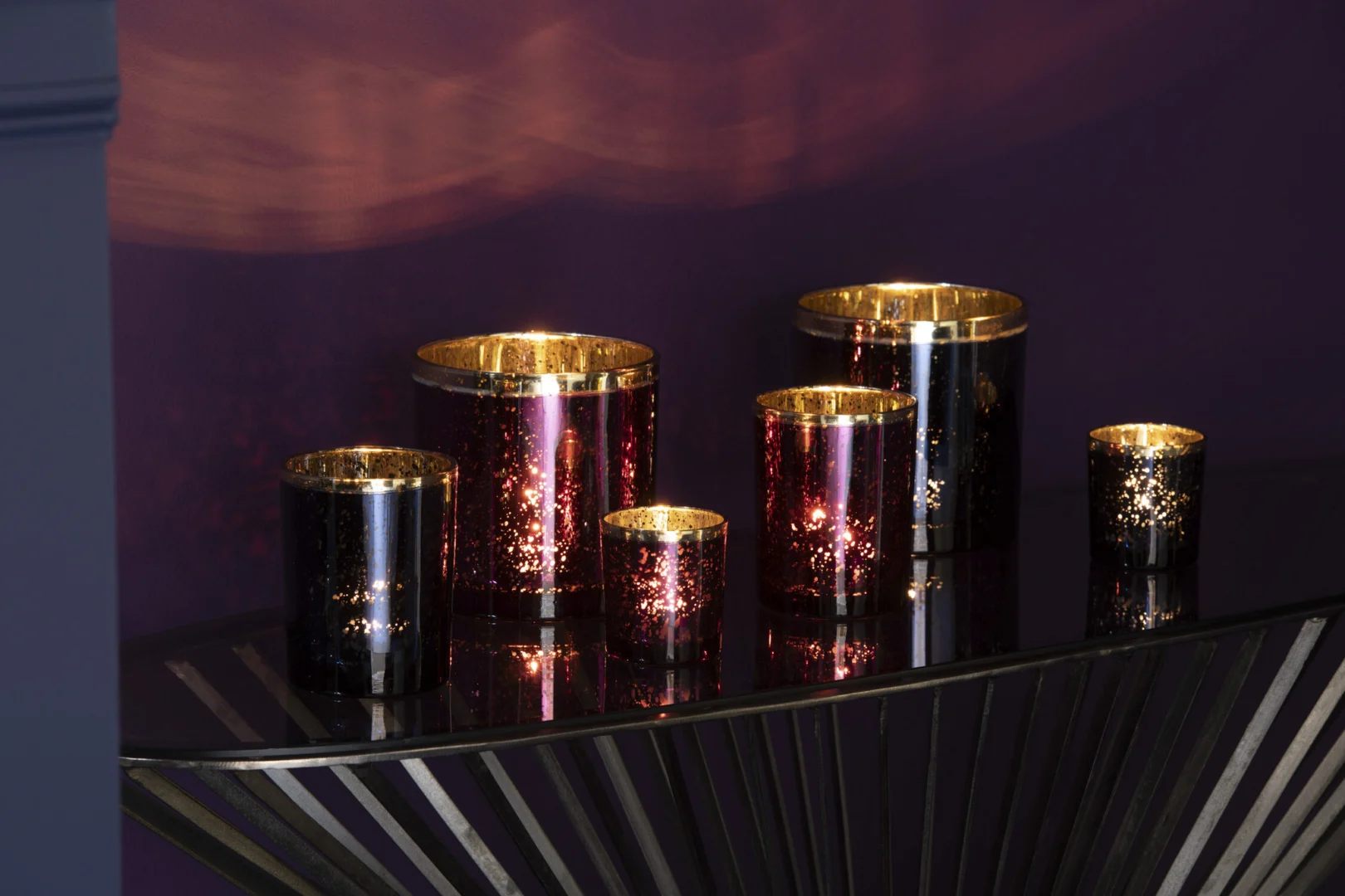 Een kleine bordeauxrode glazen kandelaar met een gouden rand, Theelichthouder met gouden rand glas bordeauxrood klein, siert een moderne consoletafel tegen een dieppaarse muur.