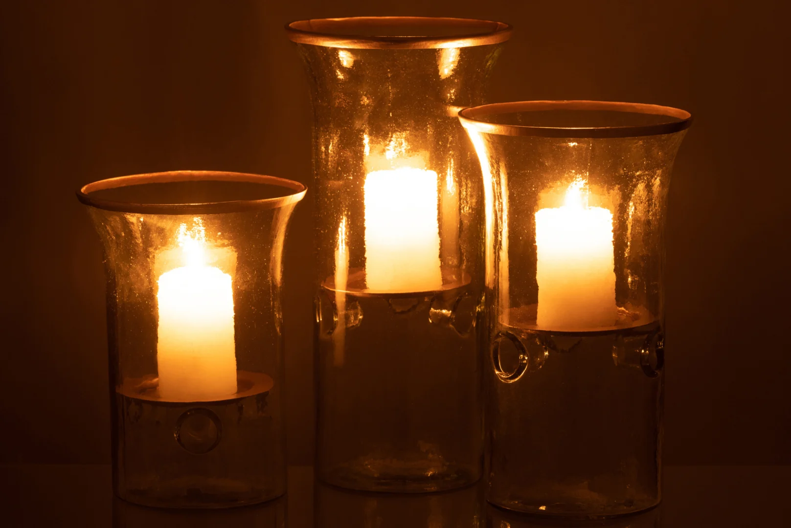 De Kaarsenhouder Glas Transparant Goud Rand Set 3 bevat drie pilaarkaarsen, waarvan de warme gloed in het transparante glas met gouden randen zacht licht en elegante reflecties werpt in een donkere omgeving.
