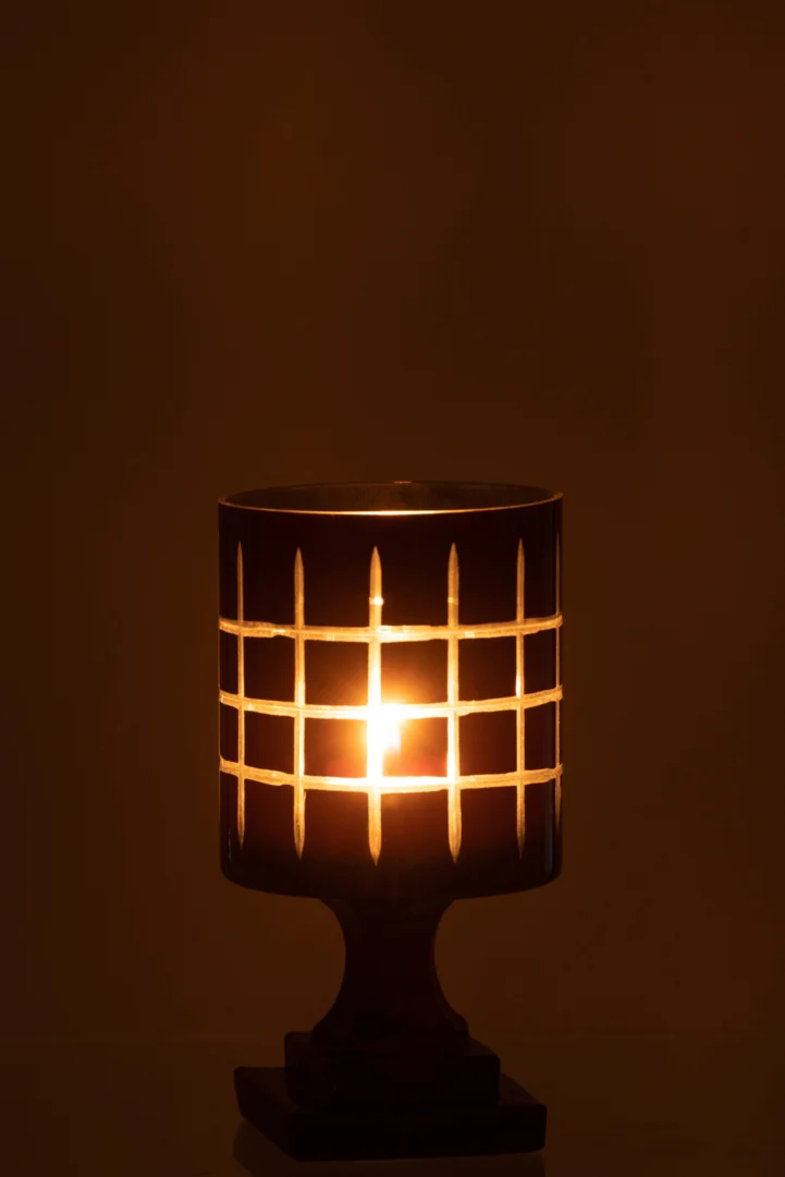De Theelichthouder Op Voet Geruit Glas Zwart gloeit met een brandende kaars binnenin en werpt warm licht en schaduwen met rasterpatronen tegen een bruine achtergrond.
