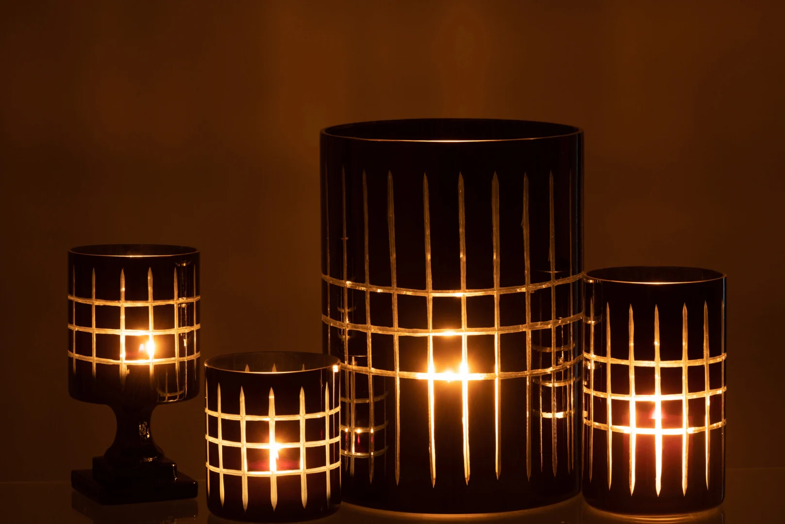 Vier Theelichthouder Op Voet Geruit Glas Zwart kandelaars met rasterpatronen stralen een warme gloed uit en werpen elegante patronen op een donker oppervlak. Hun verschillende maten geven elke ruimte een gezellige en verfijnde toets.