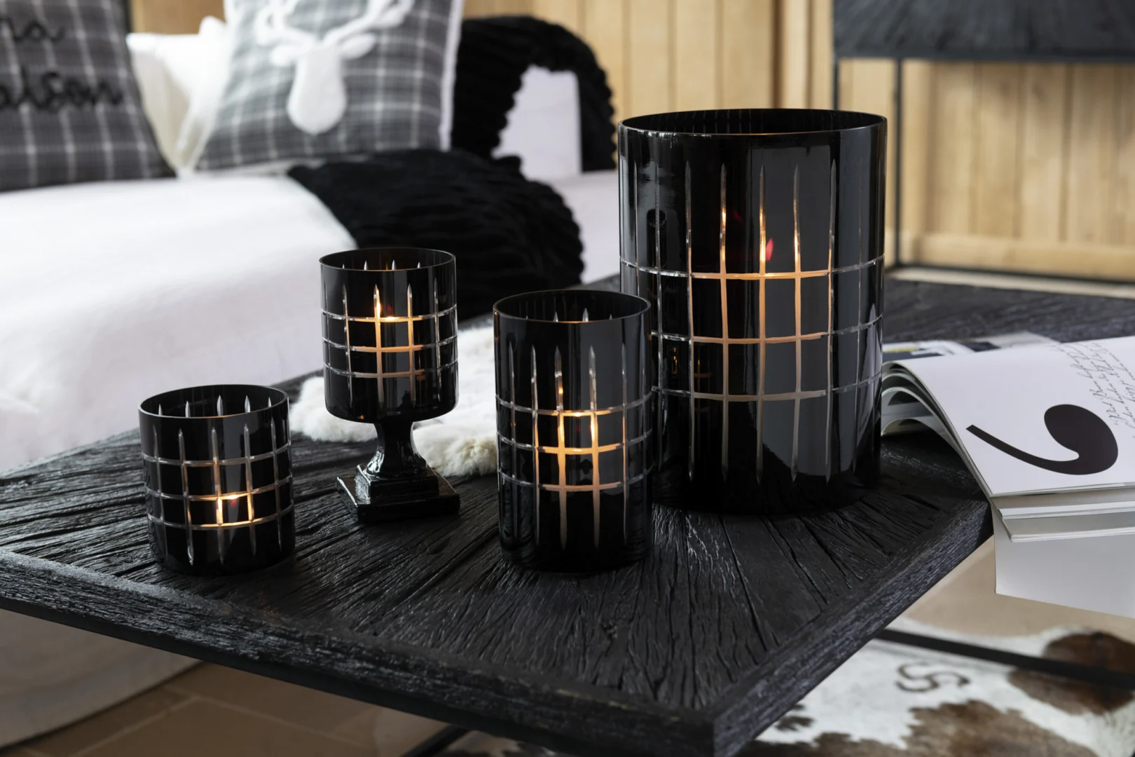 Drie Theelichthouder Op Voet Geruit Glas Zwart kandelaars, elk met een brandende kaars, staan op een donkere houten tafel naast een open tijdschrift in een gezellige moderne slaapkamer met kussens met patronen en zachte texturen op de achtergrond.