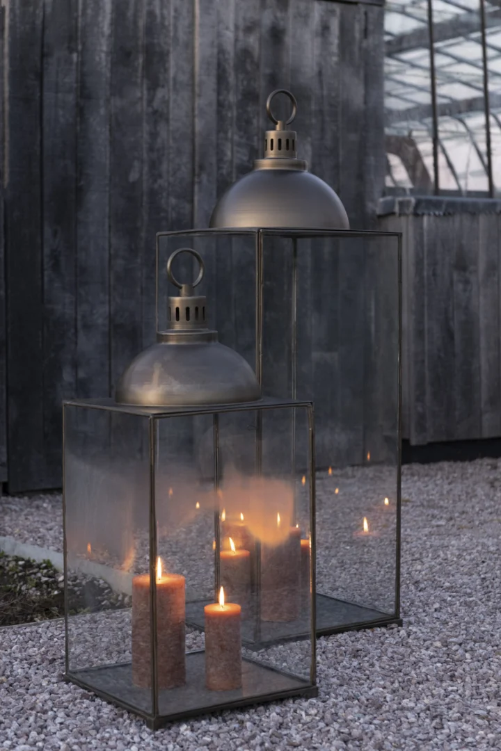 Twee grote Lantaarn Hagrid Glas IJzer Brons Grote lantaarns met brandende pilaarkaarsen staan buiten op grind, tegen een donkere houten muur en glazen kasconstructie.