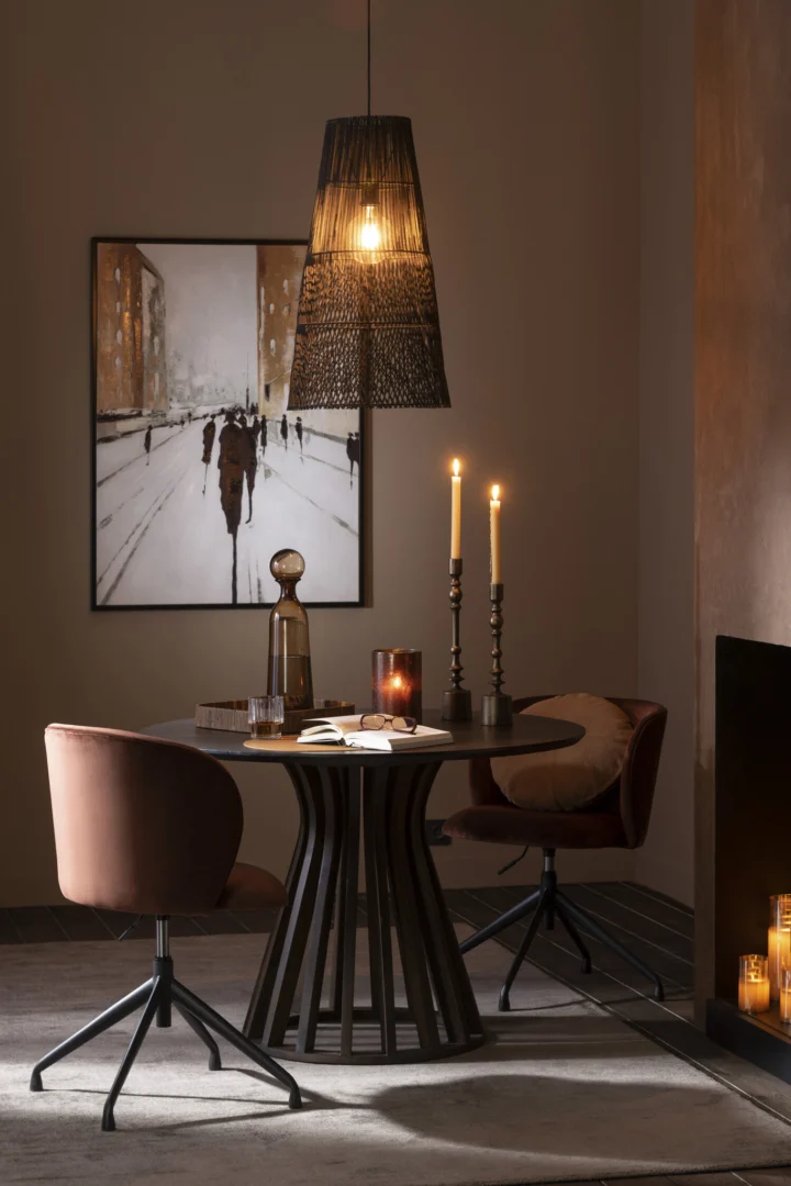 Een gezellige eethoek heeft twee fluwelen stoelen, een ronde donkere tafel met verlichte Kaarsenhouder Neuh Aluminium Brons Klein, een drankenset, moderne kunstwerken aan de muur en een geweven hanglamp die warm licht werpt in de zacht ingerichte kamer.