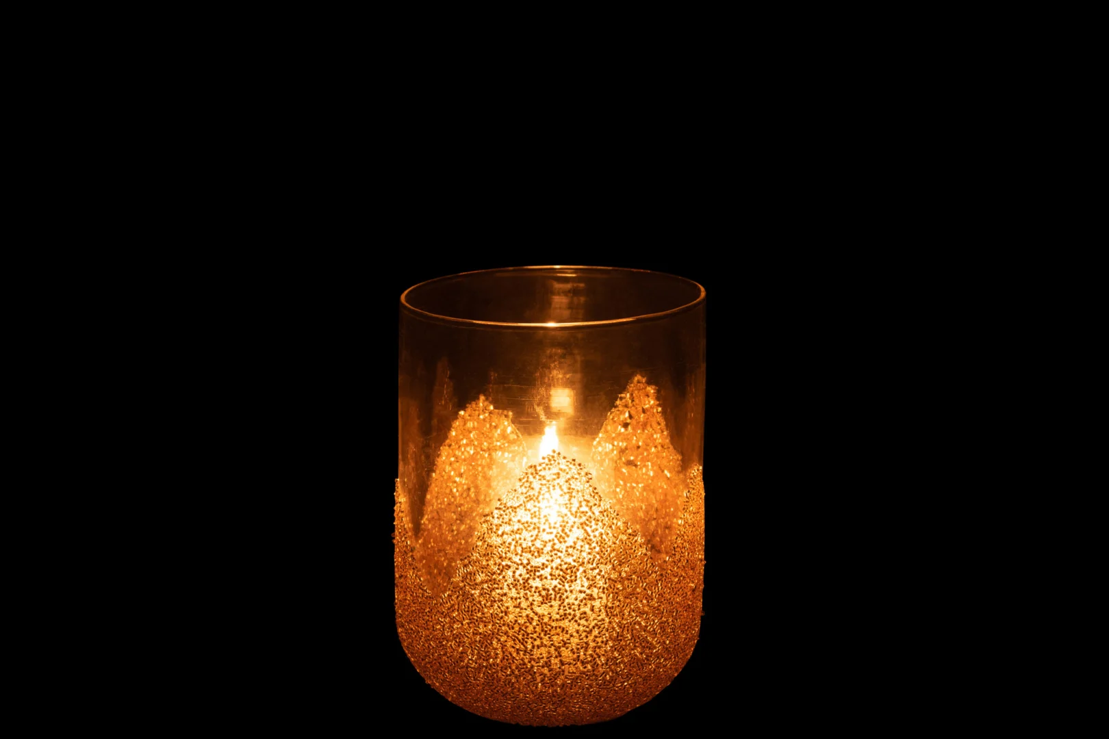 Een brandende kaars gloeit in de Kaarsenhouder Strass Glas Transparant Goud Medium, met een gestructureerd, glitterend goudpatroon tegen een zwarte achtergrond.