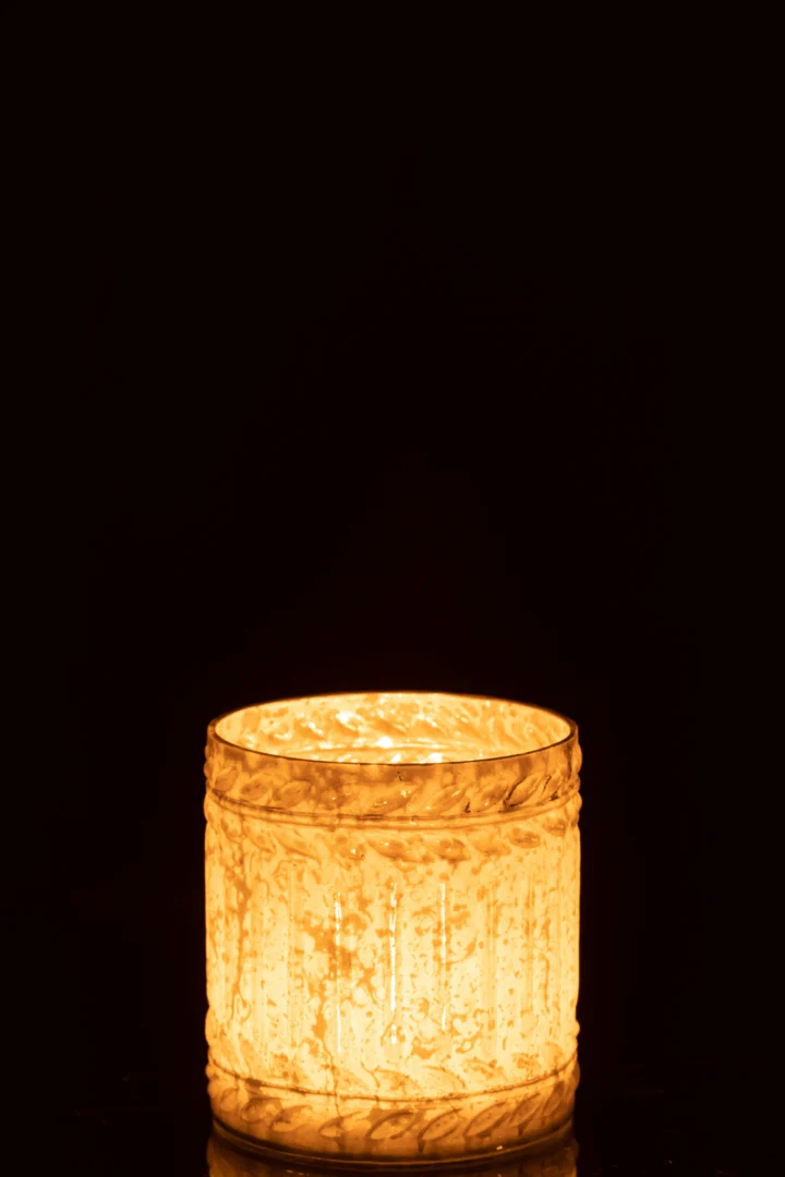 De Kaarsenhouder Ezy Glas Wit Medium kandelaar werpt een warme gloed van een enkele brandende kaars, waarbij het wit gestructureerde glas prachtig afsteekt tegen een donkere achtergrond.