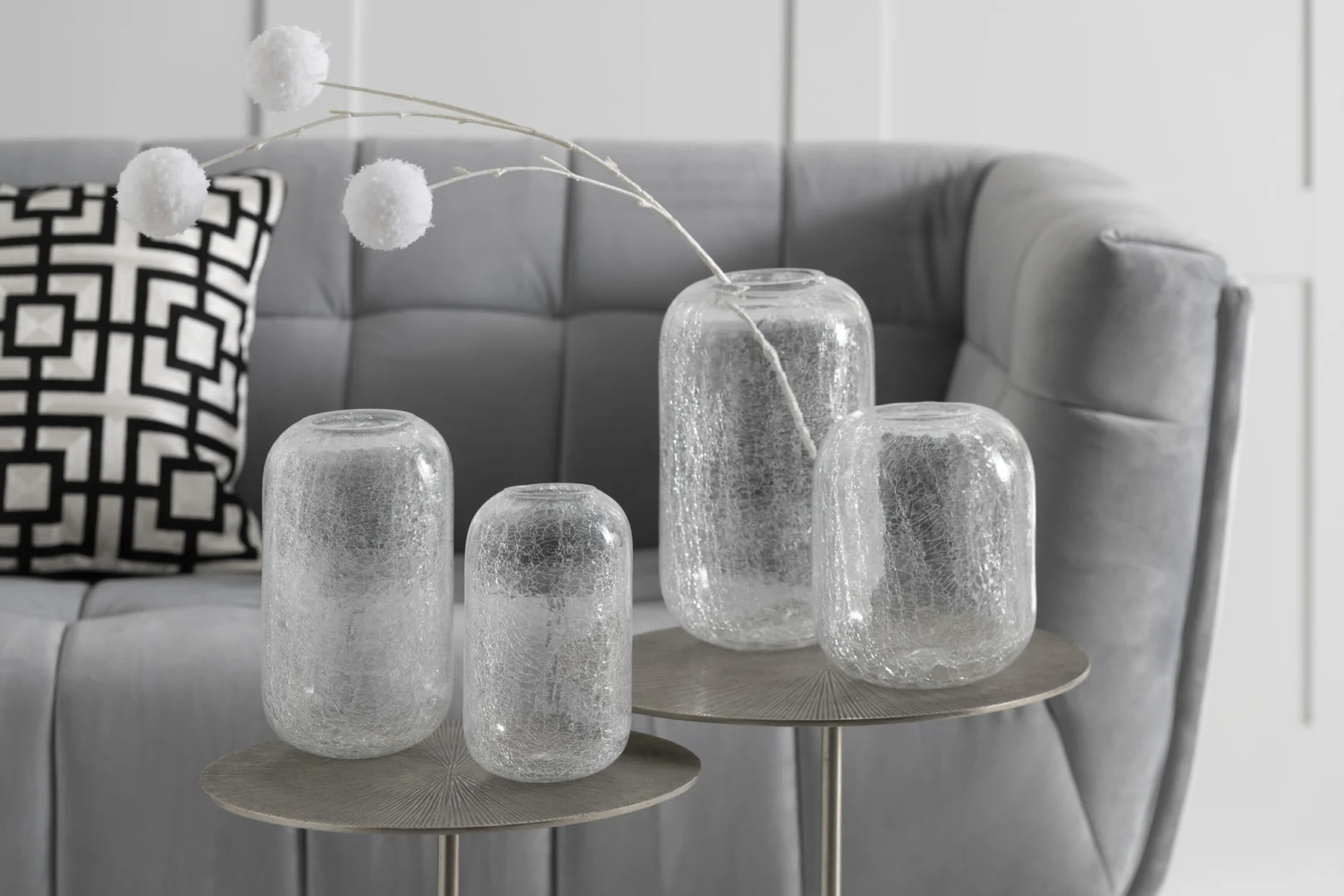 De Kaarsenhouder Orkaan Kraakglas Transparant Medium, een glazen kandelaar met textuur, staat op een ronde tafel voor een pluche grijze bank die is geaccentueerd met een geometrisch zwart-wit kussen en witte decoratieve stelen.