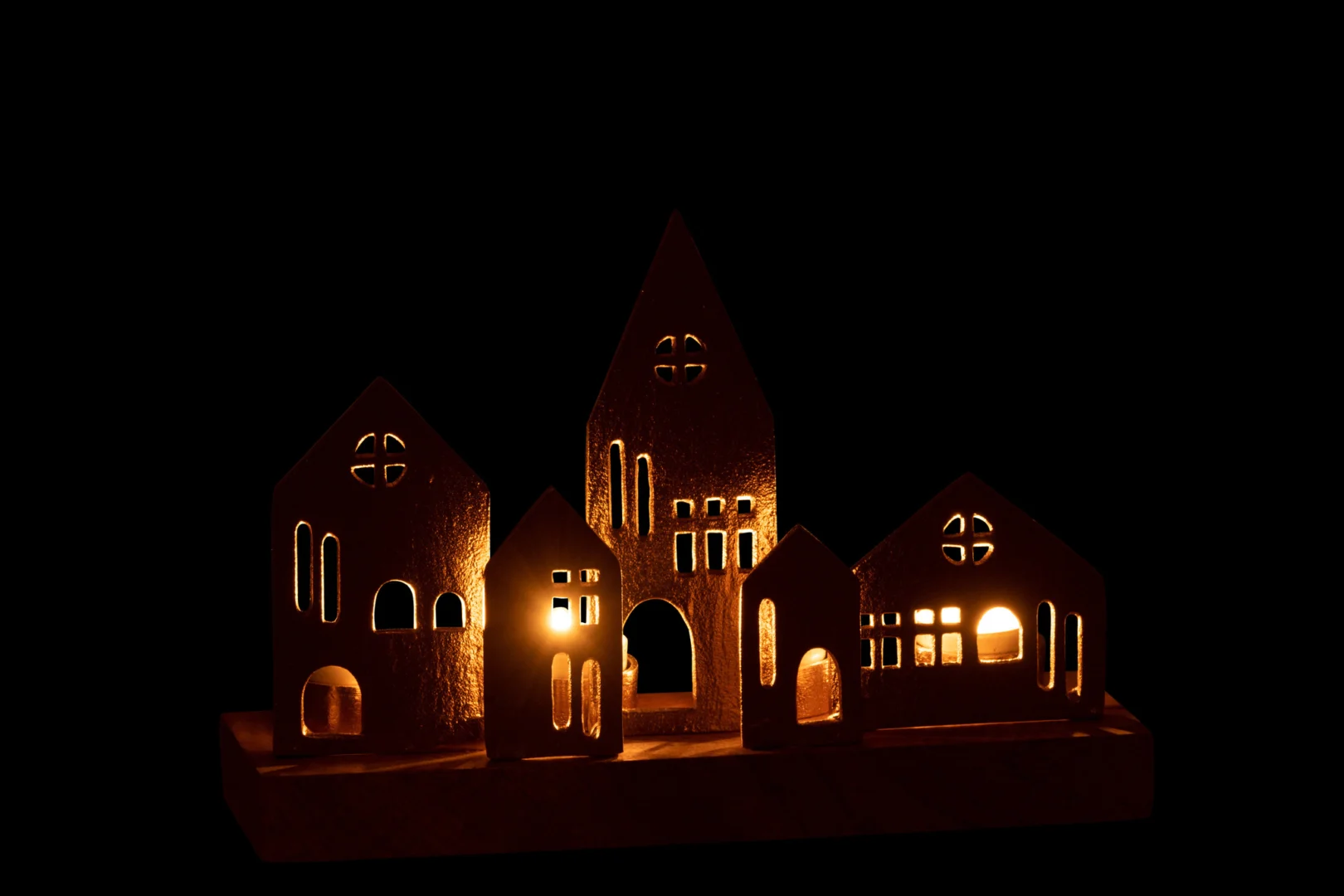 Een set Theelichthouder Huis Aluminium Hout Goud Natuurlijk: kleine, huisvormige kandelaars met uitsparingen en warmgeel licht dat binnenin gloeit, prachtig weergegeven tegen een stevige zwarte achtergrond.