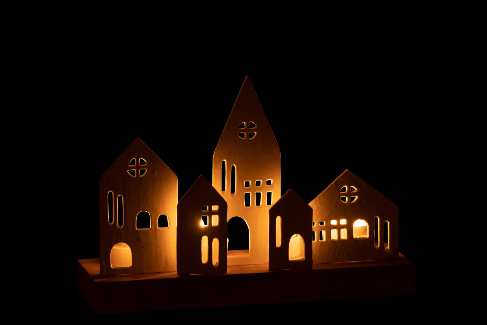 De Theelichthouder Huis Aluminium Hout Wit Natuurlijk heeft een rij decoratieve houten huizen met verlichte ramen die warm oplichten tegen een volledig zwarte achtergrond.