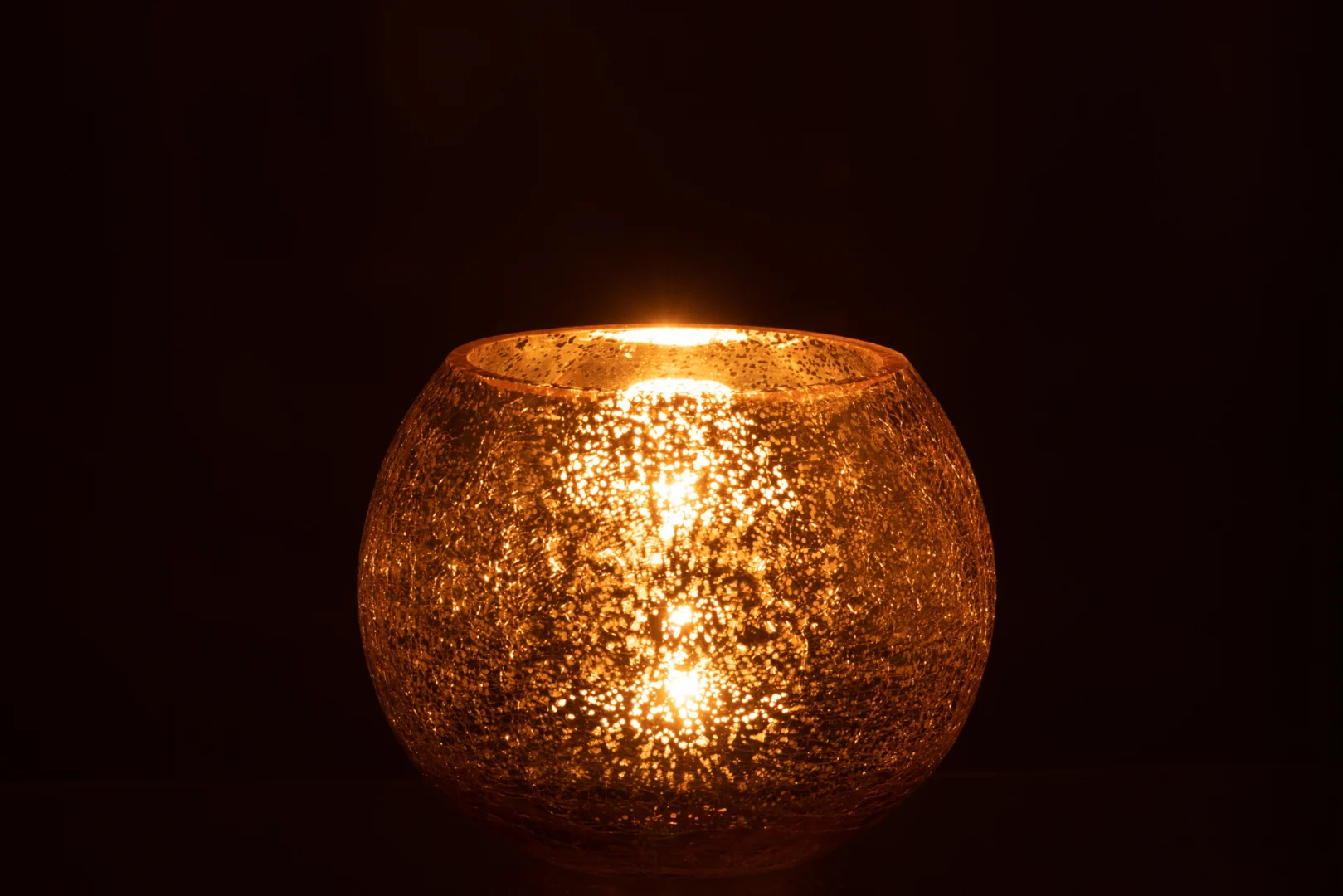 De Theelichthouder Orkaan Craquelure Glas Goud Medium heeft een gloeiende kaars in een ronde, gestructureerde glazen houder, die een warm gouden licht werpt tegen een donkere achtergrond.
