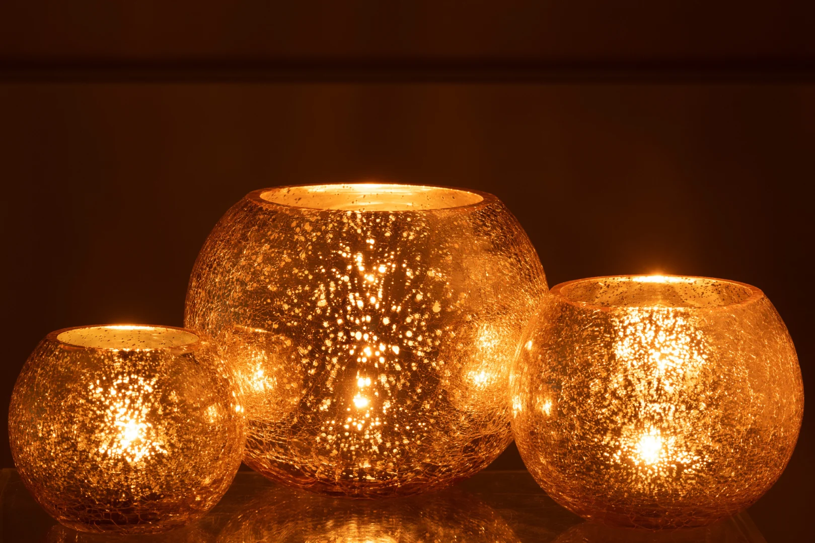 Drie grote, ronde Theelichthouder Orkaan Craquelure Glas Goud kandelaars gloeien warm met kaarsen erin en werpen fonkelende gouden reflecties in de duisternis.