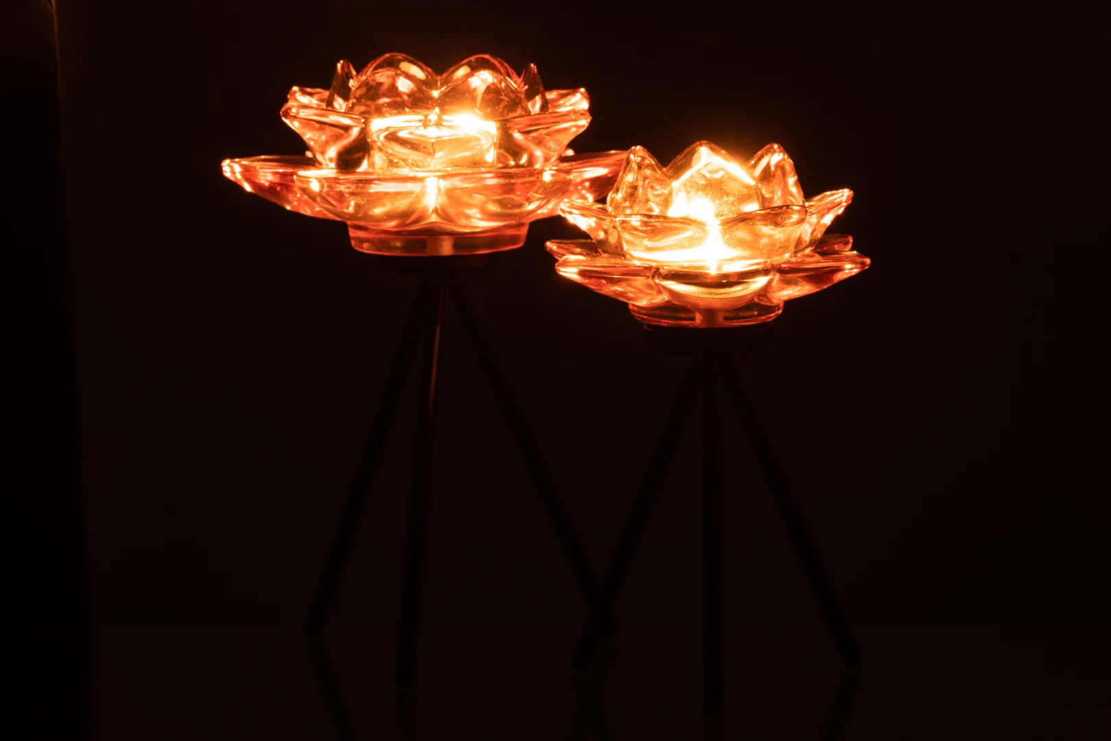 Theelichthouder Op Voet Bloem Glas Roze Groot: Twee roze glazen kandelaars in de vorm van een bloem op een hoge zwarte standaard werpen een warme, zachte gloed en verlichten de donkere achtergrond.