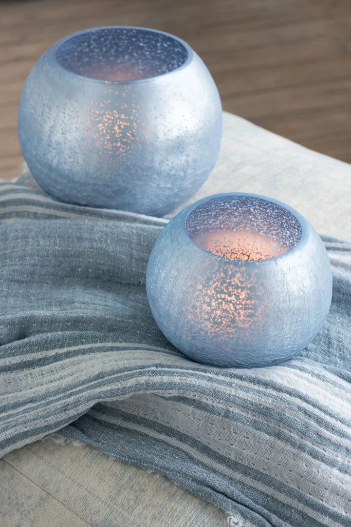 Theelichthouder Sphere Craquelure Glas Blauw Klein Set van 2 met brandende kaarsen creëren een gezellige en serene sfeer op een zacht blauw-wit gestreept kleed.