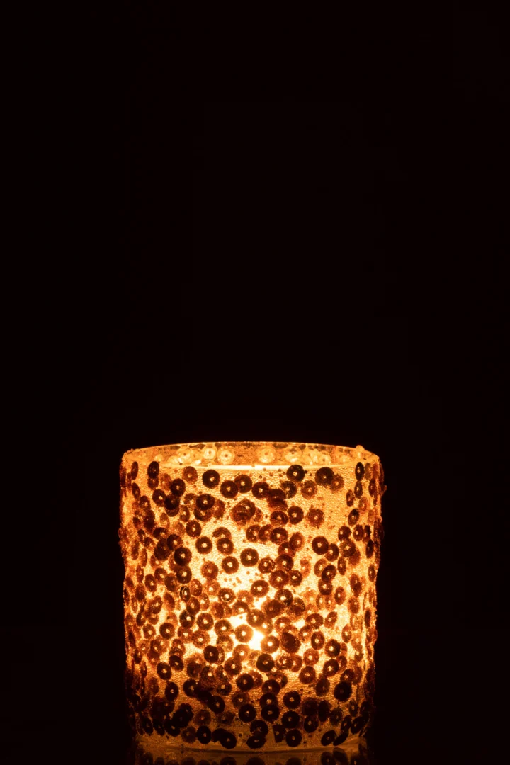De Kaarsenhouder Orkaan Sequin Glas Mat Goud Medium is een glazen kandelaar versierd met matgouden lovertjes, die een warme, zachte gloed verspreiden wanneer ze worden aangestoken tegen een donkere achtergrond.