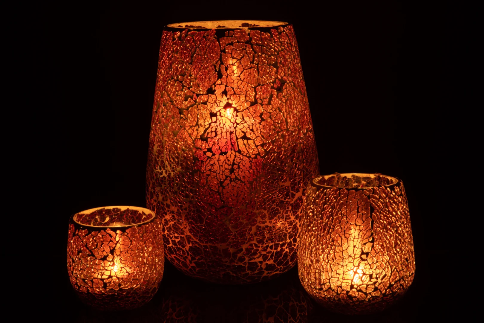 De Kaarsenhouder Ali Glas Bruin Groot staat hoog in het midden, met zijn gebarsten mozaïekglas dat warm gloeit met zacht goudoranje licht, geflankeerd door twee kleinere houders die een gezellige sfeer creëren in het donker.