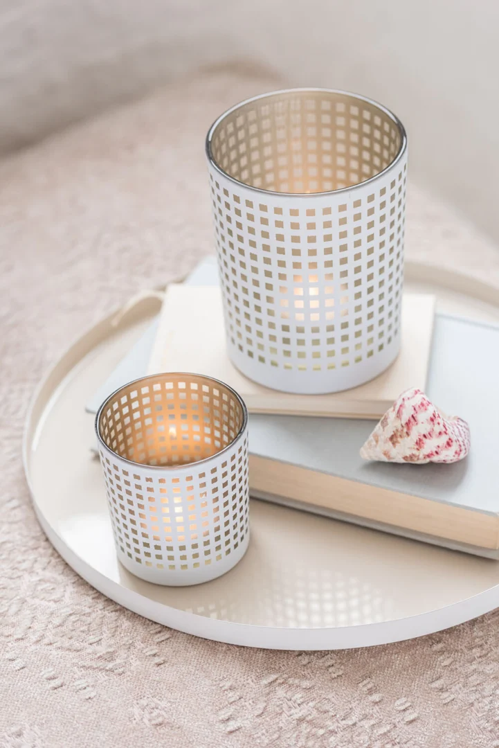 Twee Theelichthouder Patroon Glas Wit Beige kandelaars met brandende kaarsen staan op een rond wit dienblad met gestapelde boeken en een schelp, uitgestald op een gestructureerd oppervlak van lichte stof.