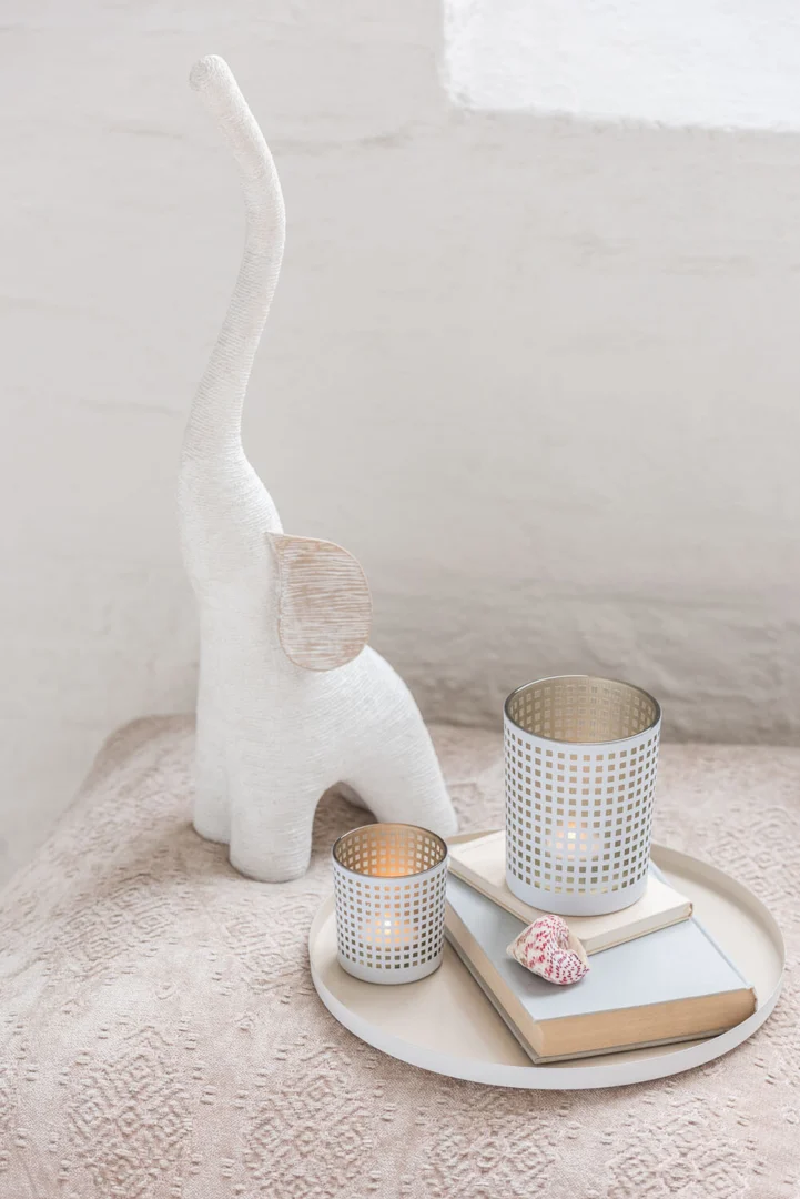 Een witte decoratieve olifant staat naast een rond dienblad met daarop Theelichthouder Patroon Glas Wit Beige Set 2 met brandende kaarsen, twee gestapelde boeken en een kleine roze schelp op een beige gestructureerd oppervlak.