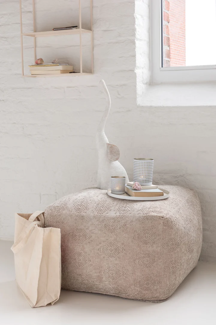 Een set Theelichthouder Patroon Glas Wit Beige ligt op een knusse poef bij een witte bakstenen muur en raam en creëert een warme, minimalistische sfeer. Een beige tas maakt de serene look compleet.