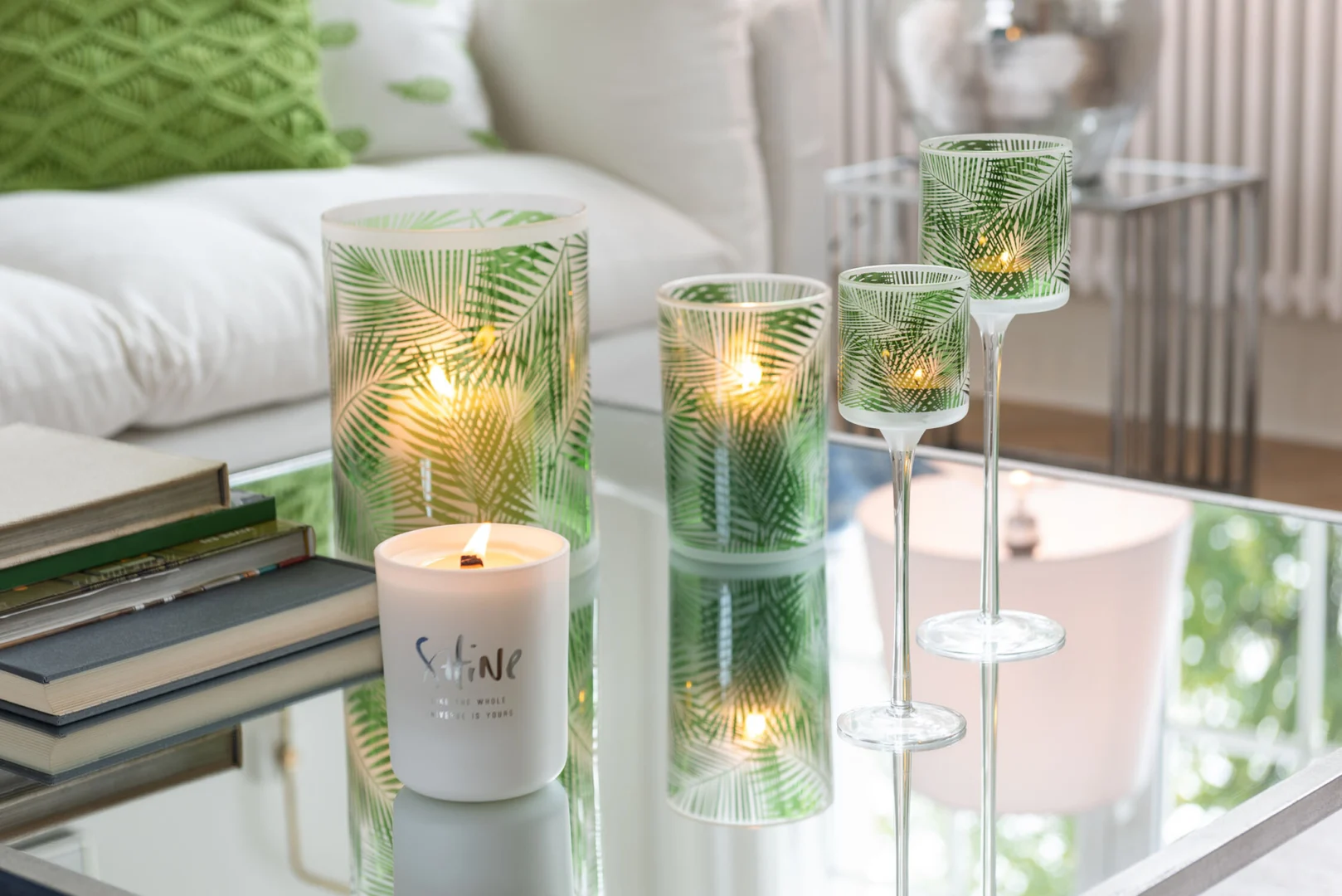 Een witte kaars met het label "Shine" en drie Kaarsenhouder Orkaan Tropisch Glas Groen Groot kandelaars rusten op een spiegelende tafel in een moderne woonkamer met een witte bank en groene kussens.
