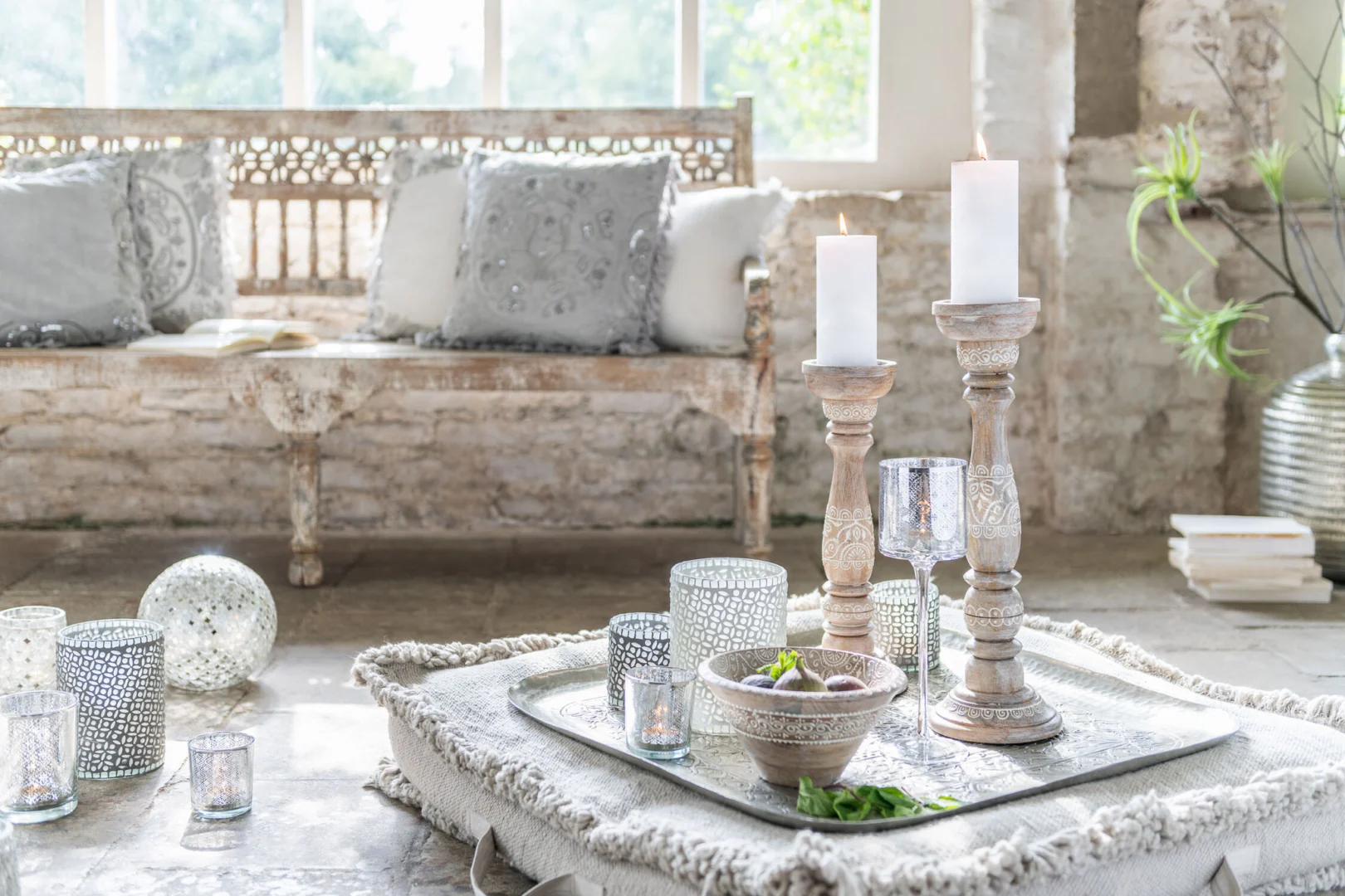 Een gezellige, rustieke woonkamer met een verweerde houten bank, decoratieve kussens, verlichte Theelichthouder Op Voet Oosters Glas Zilver Groot kandelaars, een dienblad met fruit en zacht natuurlijk licht dat door de grote ramen naar binnen valt.