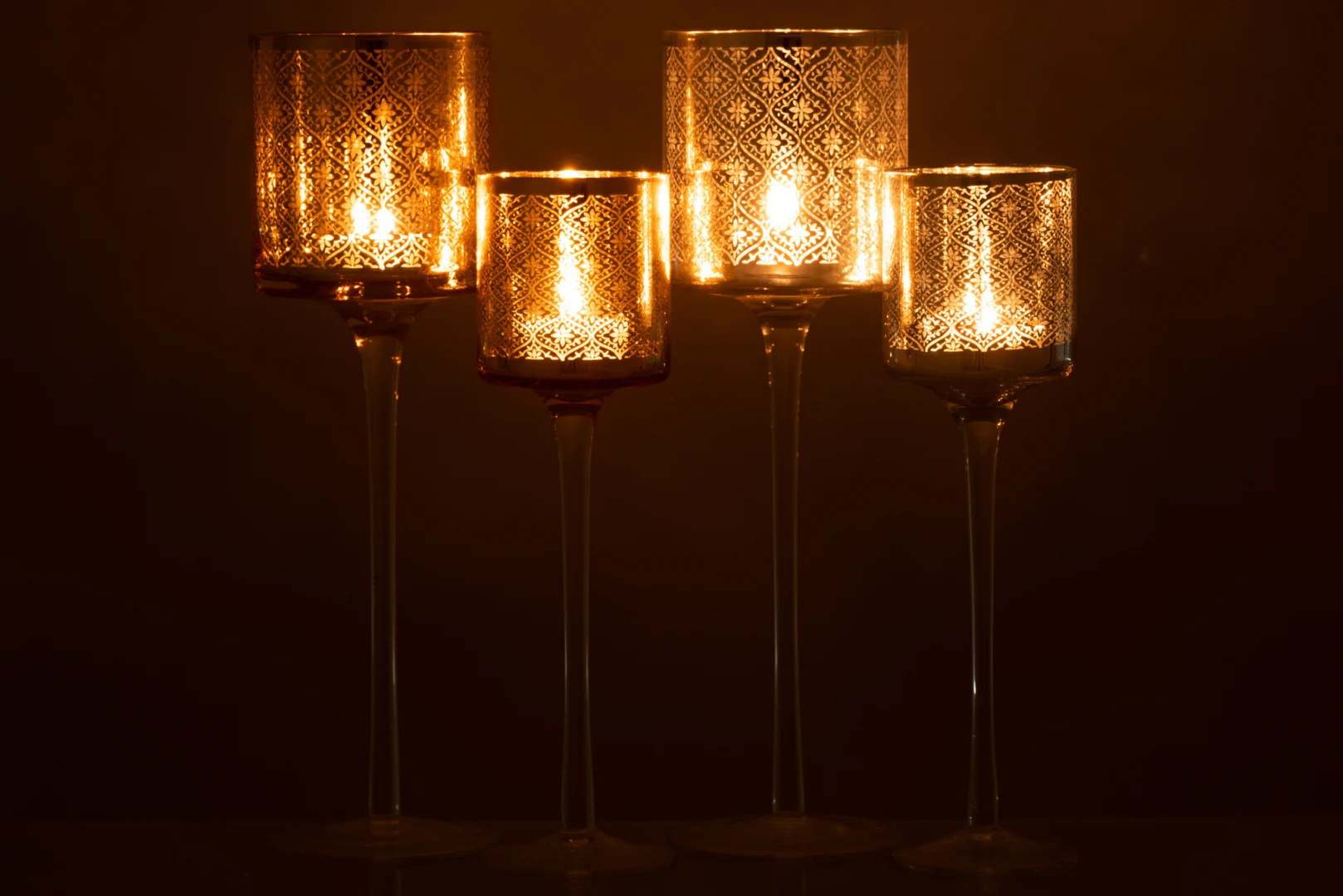 Vier gedessineerde Theelichthouder Op Voet Oosters Glas Zilver Groot kandelaars op hoge stelen branden en werpen een warme gloed tegen een donkere achtergrond. Hun ingewikkelde ontwerpen creëren een elegante, decoratieve sfeer.