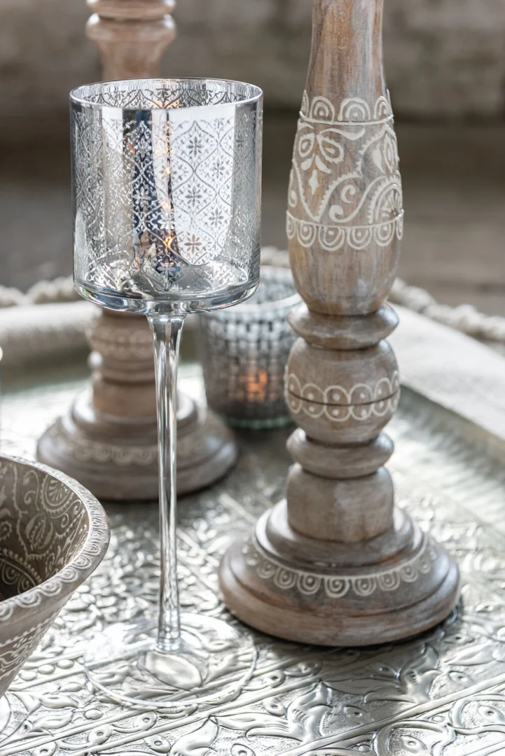 De Theelichthouder Op Voet Oosters Glas Zilver Groot staat hoog naast houten kandelaars en een gedessineerde schaal, allemaal gerangschikt op een sierlijk zilveren dienblad met ingewikkelde ontwerpen, terwijl een kleine kaars op de achtergrond gloeit.